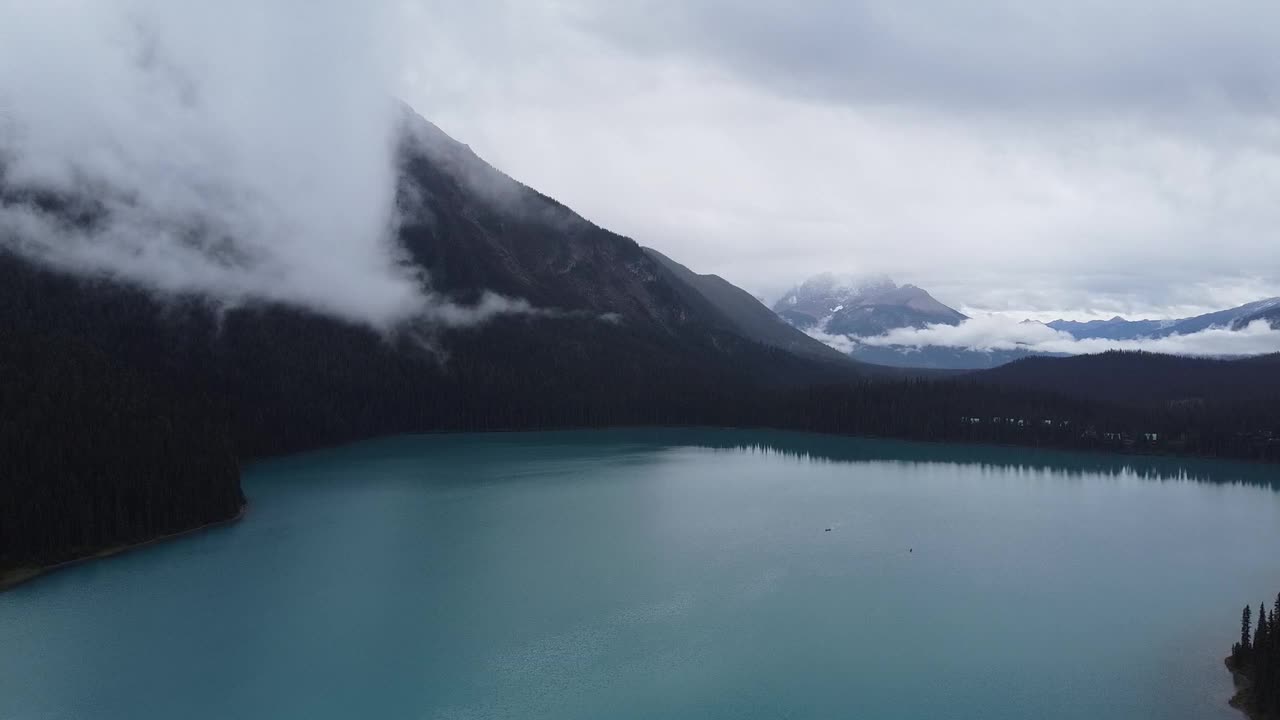 Aerial drone shot of calm turquoise lake bordered by dark forest and towering mist-covered mountains under overcast sky in Canada, capturing dramatic natural scenery and remote mountain wilderness