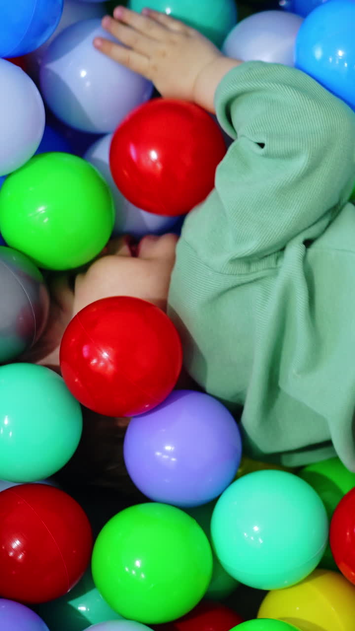 Happy baby boy playing inside the dry soft pool. Kid turns around and picks one ball. Top view. Vertical video