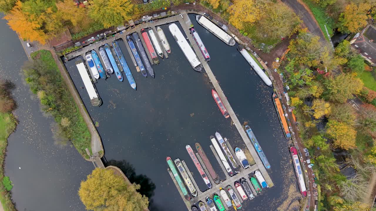 High drone orbit over Harlow Marina, Essex. The top-down view shows colourful narrowboats surrounded by autumn trees. The still water reflects leaves and debris, creating a calm seasonal aerial scene