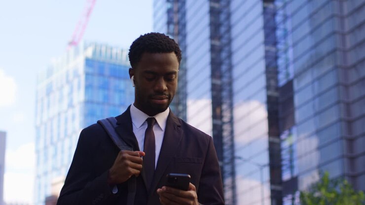 Young Businessman Wearing Wireless Earbuds Streaming Music Or Podcast From Mobile Phone Walking To Work In Offices In The Financial District Of The City Of London UK Shot In Real Time