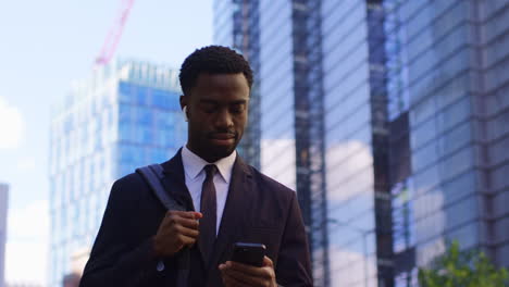 young businessman wearing wireless earbuds streaming music or podcast from mobile phone walking to work in offices in the financial district of the city of london uk shot in real time