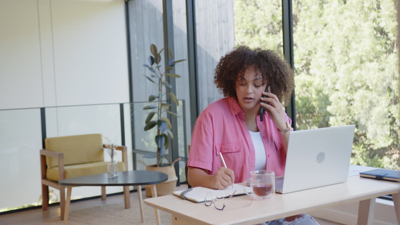 Talking on phone, woman working on laptop and writing in notepad at home office
