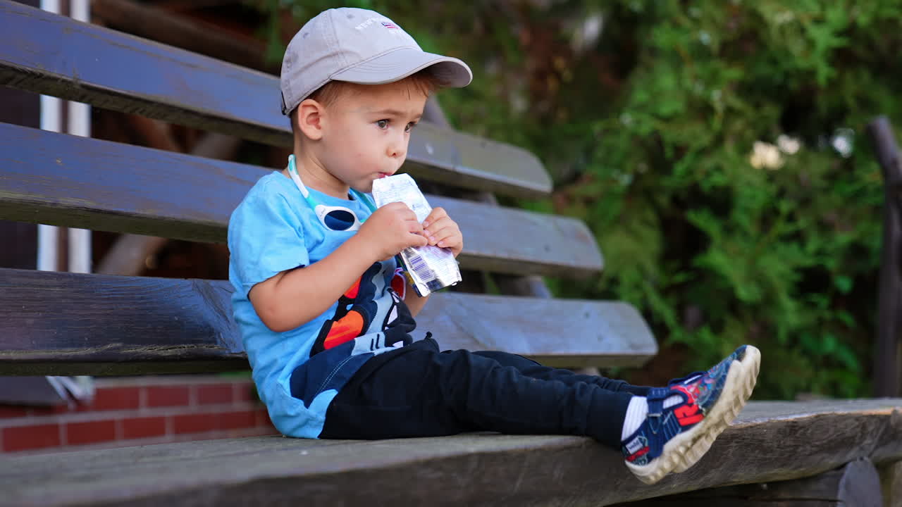 Two year old toddler sitting on the bench outdoors in summer. Baby boy drinking from a doy pack.