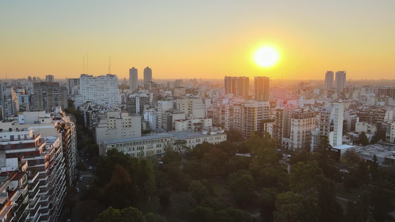 Buenos Aires cityscape at sunset, tall buildings in the background and Parque Rivadavia's green canopy in the foreground. Slow aerial descend