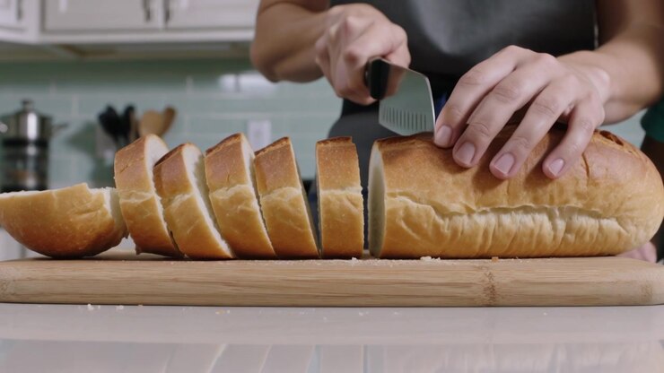 Slicing adult in grey apron with serrated knife on cutting board in kitchen, child steadying bread