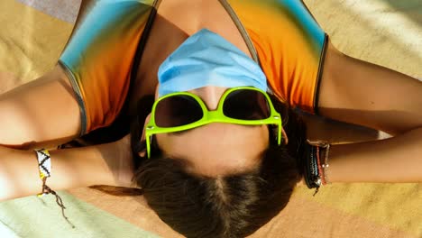 covid de verano, chica con mascarilla se pone gafas de sol, acostada sobre una toalla colorida
