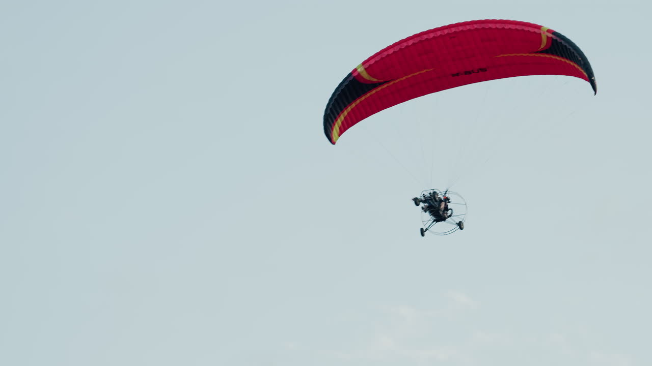 upward view of paramotor pilot ascending beneath red canopy against pale cloudy sky showcasing motorized frame silhouette and open air adventure above misty horizon