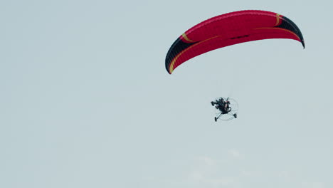 upward view of paramotor pilot ascending beneath red canopy against pale cloudy sky showcasing motorized frame silhouette and open air adventure above misty horizon