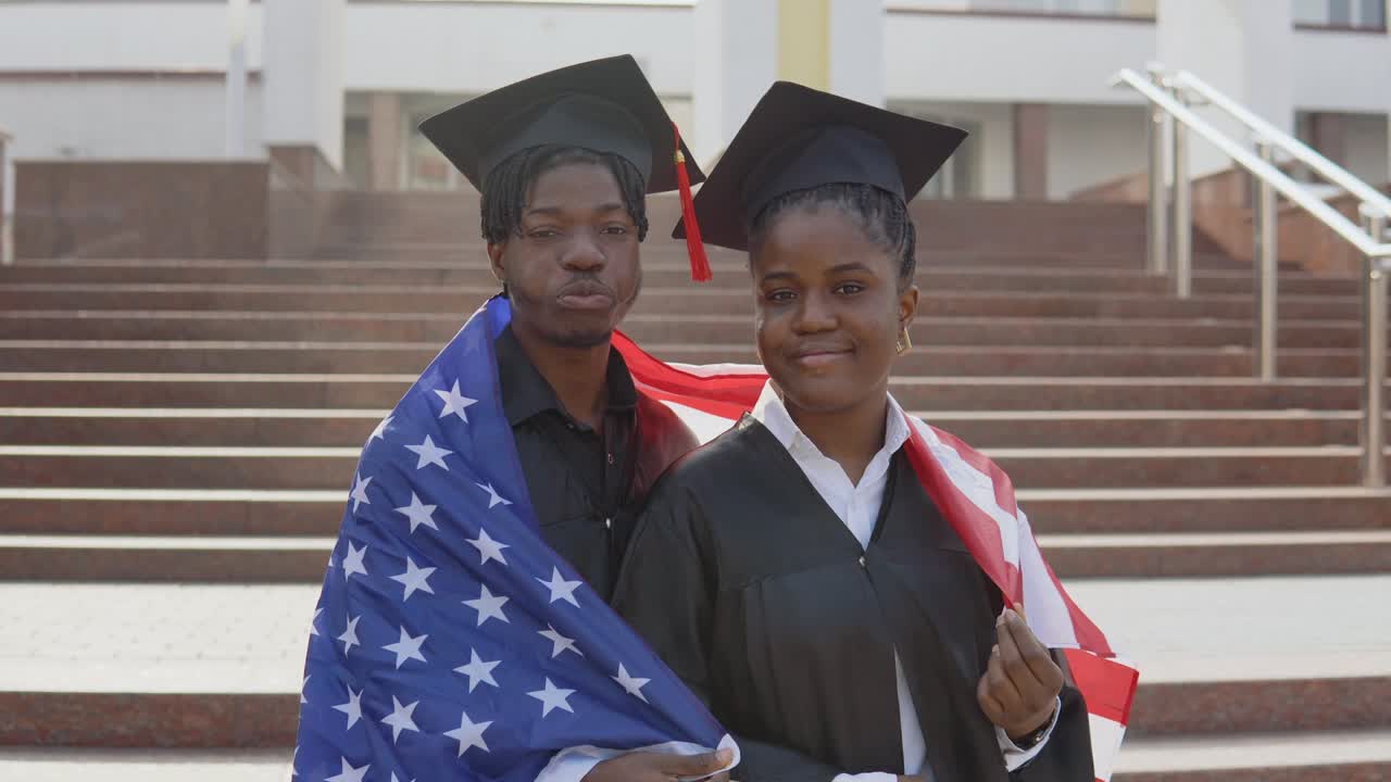 un hombre y una mujer afroamericanos se paran uno al lado del otro frente a la cámara con túnicas negras y sombreros cuadrados de estudiantes graduados con la bandera de los estados unidos en los hombros