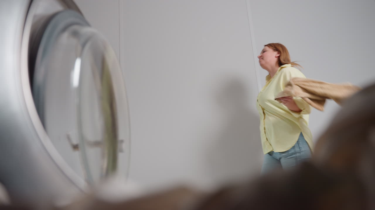 Vista dall'interno del cestello di una lavatrice che mostra una mamma felice che lancia dei vestiti mentre passa sorridendo; riflesso tenue sul bordo di vetro, lavanderia luminosa, atmosfera moderna, prospettiva dalla porta con bordo