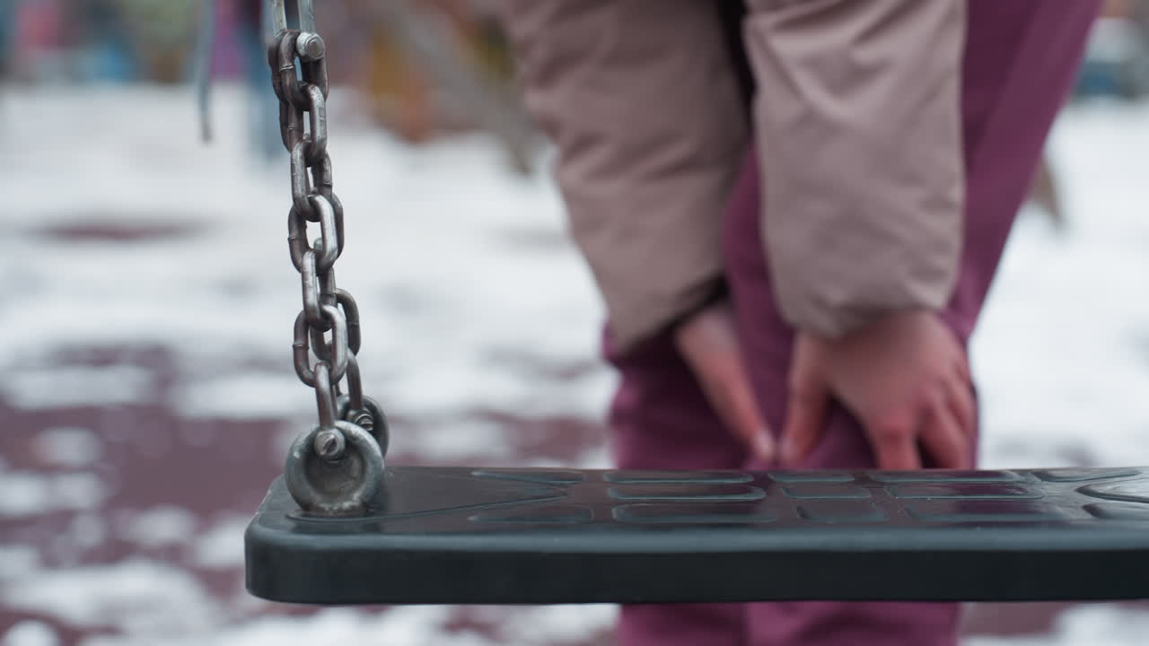 primer plano de swing con una persona en traje de invierno frotando la rodilla izquierda, posiblemente con dolor, swing en primer plano con fondo borroso que muestra a alguien balanceándose en la distancia