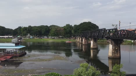 Drone-footage-revealing-the-side-of-the-River-Kwai-Memorial-Bridge-and-the-other-side-of-the-river-houses-built-around-the-picturesque-historic-landscape,-Kanchanaburi,-Thailand