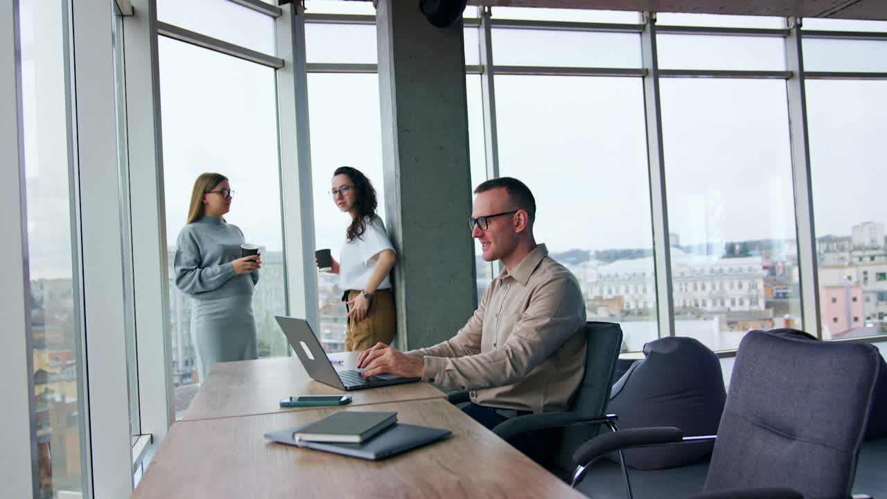 Man wearing eyeglasses types on his laptop. Female colleagues chat cheerfully standing at the window with coffee cups.