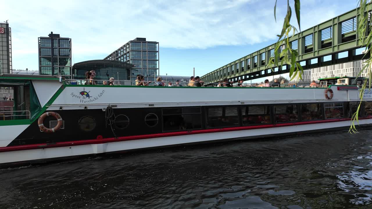 A river cruise boat navigates through Berlin, passing by lush trees and modern architecture. The scene captures the blend of nature and urban life.