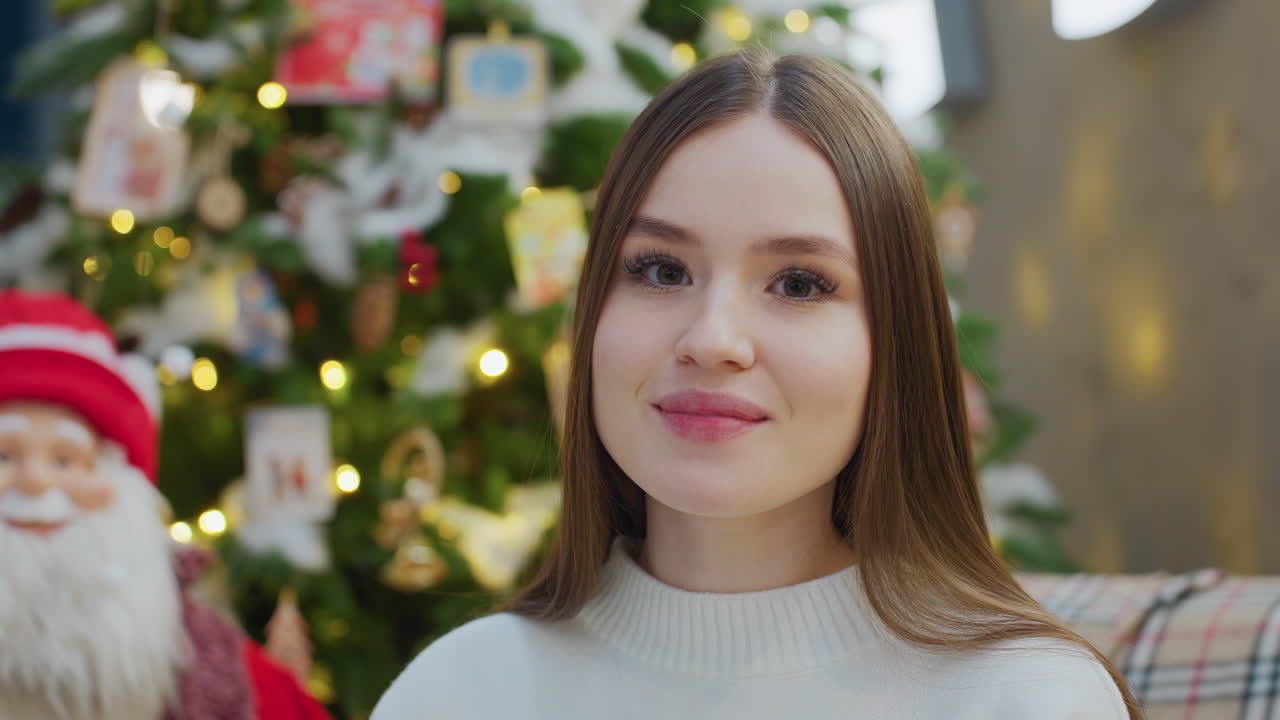 Elegant lady smiling warmly while sitting near beautifully decorated Christmas tree with glowing lights and colorful ornaments, creating a festive and cheerful holiday atmosphere