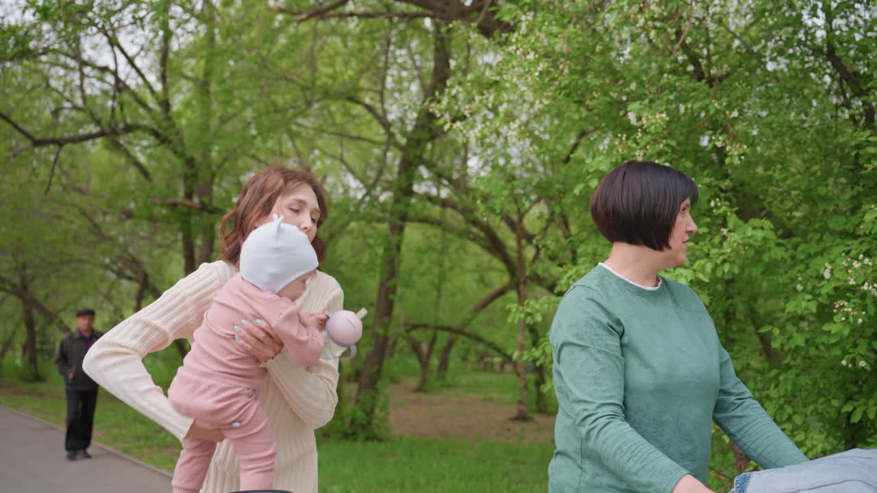 Women Chatting Peacefully, Gentle Conversation During Outdoor Walk, Two Ladies Converse Softly Beneath Leafy Trees, Two Females Converse Harmoniously While Cradling Child During Serene Park Walk