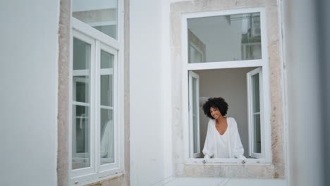 Chica-Doméstica-Asomando-Por-La-Ventana-Por-La-Mañana-En-El-Interior.-Señora-Africana-Mirando-La-Cámara-Sola