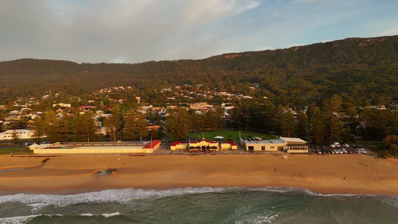 Aerial pullback over Thirroul Beach surf club with sand and waves nearby under sunny skies
