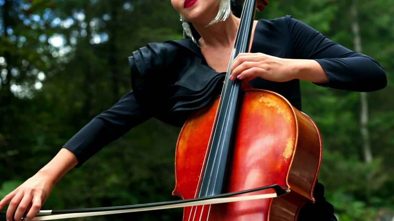 Cello in woman's hands outdoors. Beautiful woman with earrings and black dress playing the musical instrument on nature background.