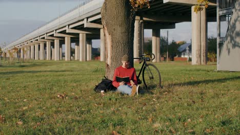 man reading a book in a park underneath a bridge