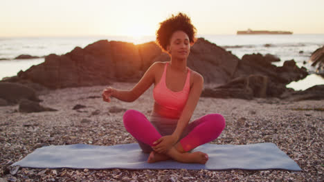 african american woman practicing yoga and meditating on the rocks near the sea during sunset