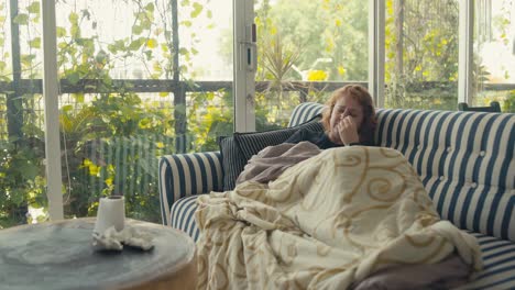 Young-sick-caucasian-woman-suffering-with-flu,-A-lady-sitting-alone-on-couch-sneezing-under-the-blanket