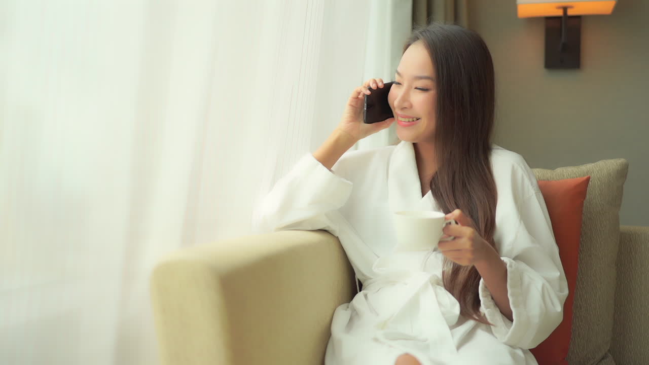 Relax at home. Chinese girl talking on the phone and drinking coffee wearing bathrobe
