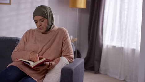 Muslim-Woman-Wearing-Hijab-Sitting-On-Sofa-At-Home-Reading-Or-Studying-The-Quran-2