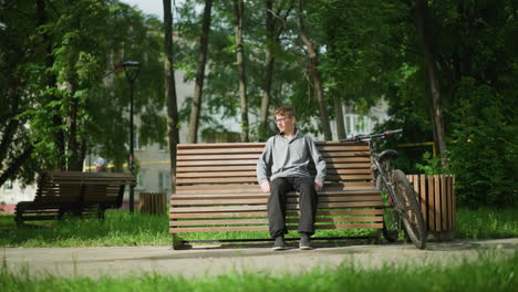 boy wearing glasses, gray top, and black trousers sits on wooden bench outdoors, stretching and adjusting himself while resting, bicycle parked beside him, someone seated in background