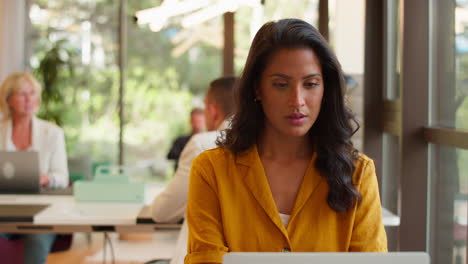 Mature-Businesswoman-Working-At-Desk-In-Open-Plan-Office-Looking-At-Watch