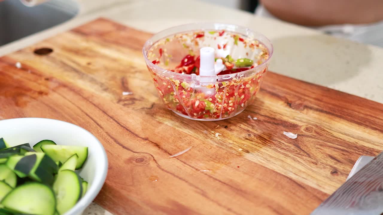 Hands use a manual chopper to finely chop garlic and chili, surrounded by fresh ingredients on a wooden board