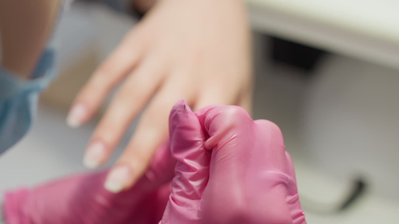 Close up slightly blurred view of nail technician in pink gloves gently applying product to client hand using pink brush during manicure session under salon lighting on white surface