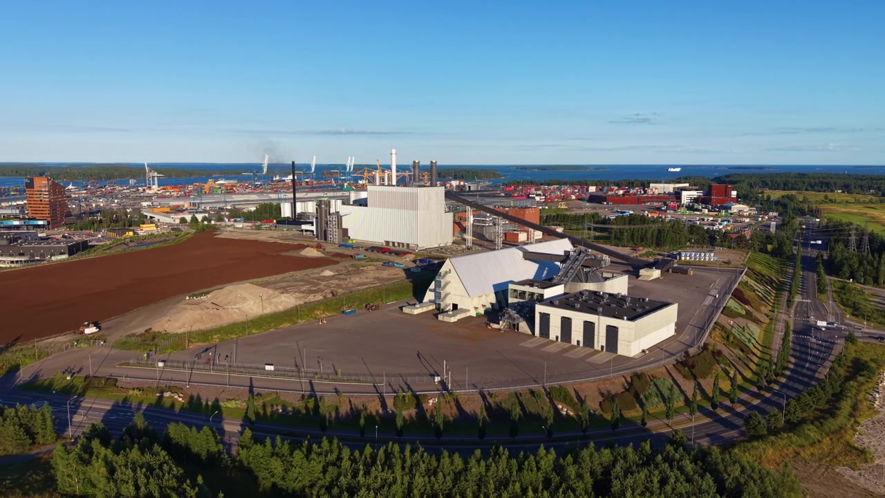 Oversiktsbilde av et biomassefyrt fjernvarmeanlegg ved siden av Vuosaari containerhavn i Helsingfors, Finland, omgitt av skog, åpent landskap og Østersjøkystlinje.