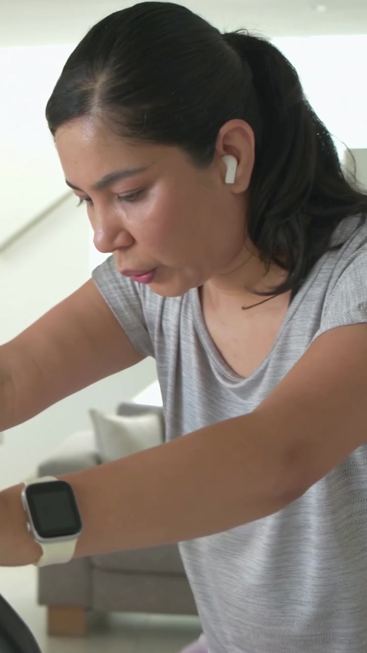 Vertical video: Pedaling woman in lilac leggings monitoring smartwatch during home bike session