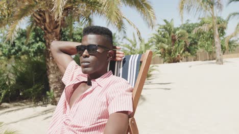 african american man sitting on deckchair wearing sunglasses, relaxing on sunny beach, slow motion