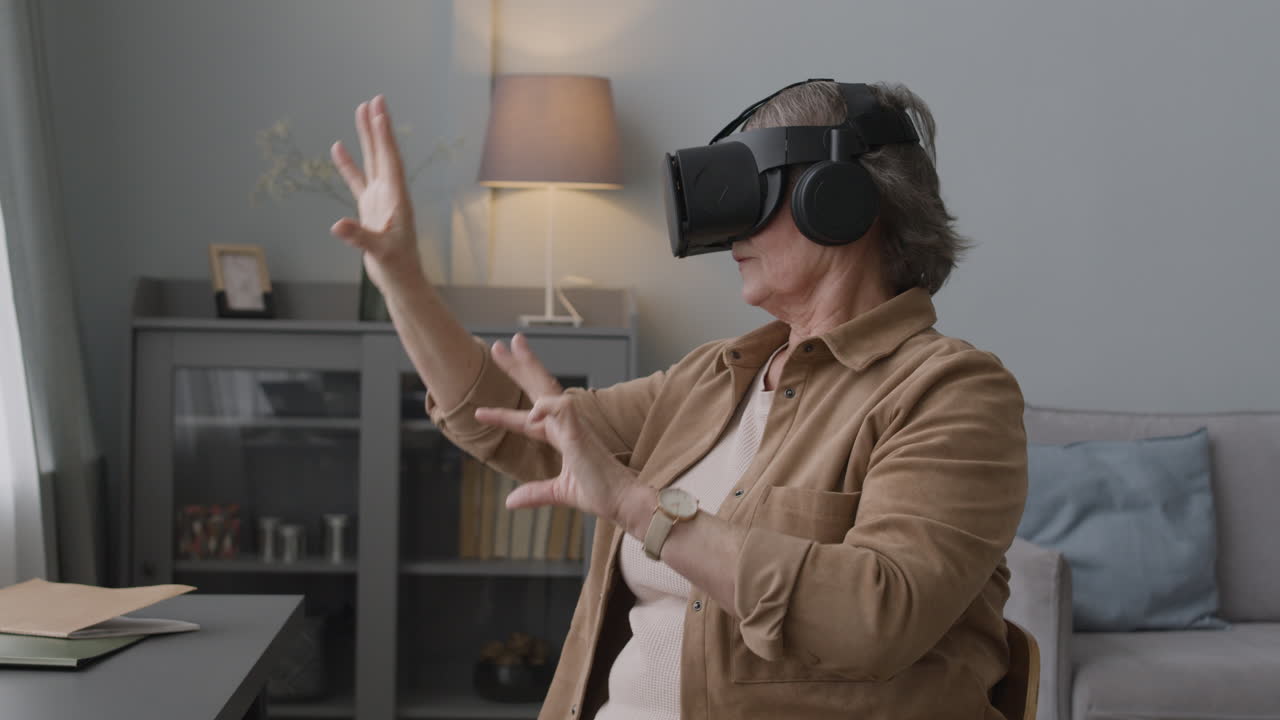 пожилая женщина использует очки гарнитуры виртуальной реальности и двигает руками, сидя на стуле в современной гостиной 2