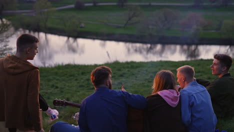group of young people sitting on the grass hear campfire and singing songs. friends enjoy the sunset and view of the river in front of them.