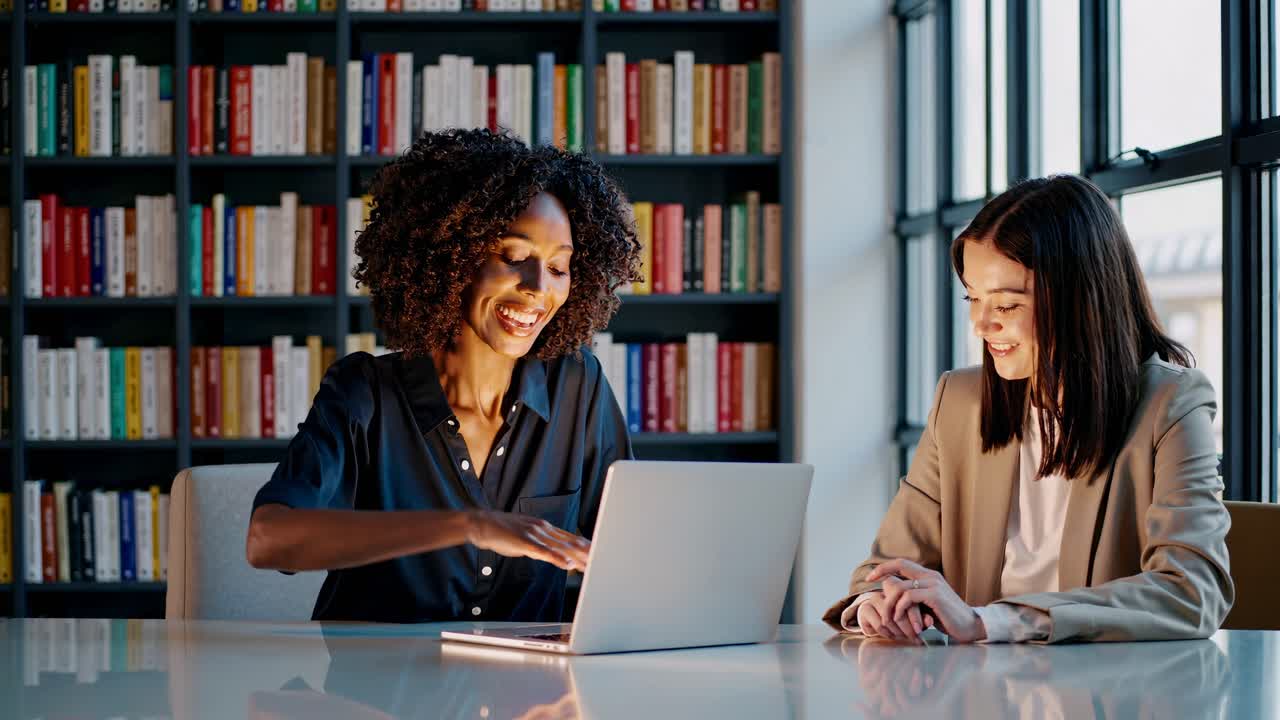 Two professionals in a library setting, smiling and working on a laptop