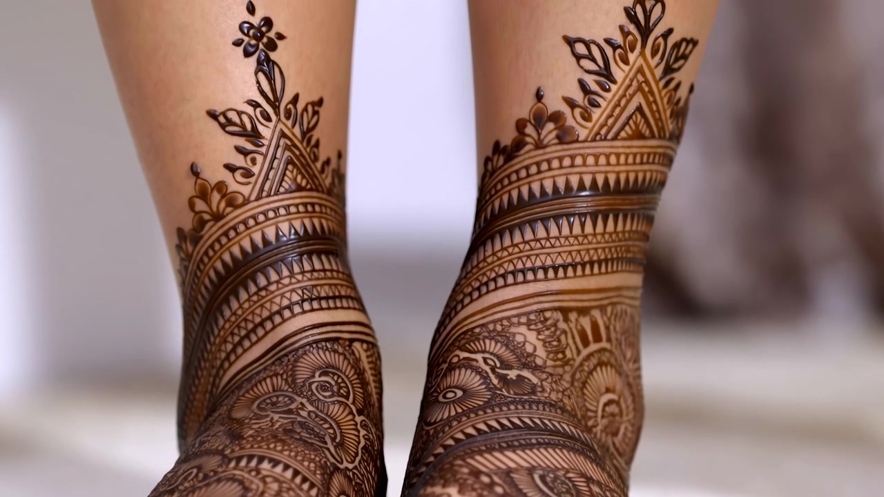 Vista ravvicinata di due piedi e parte inferiore delle gambe coperti da intricati disegni di henné marrone scuro, con motivi floreali, geometrici, simili a pizzi e ispirati agli occhi, tonalità della pelle calde, illuminazione soffusa e sfondo sfocato.