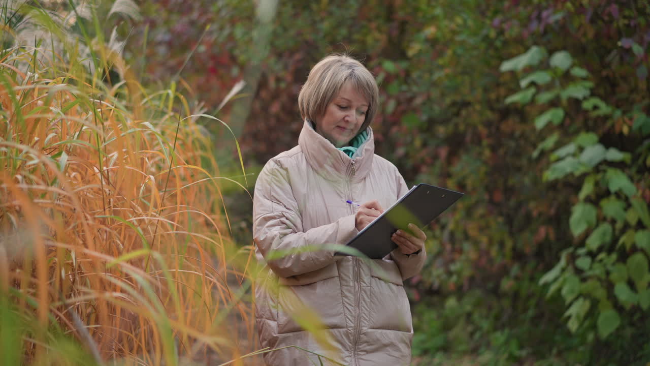 middle aged woman bundled in light coat stands by tall golden bush writing notes on clipboard, calmly observing and documenting plant details while surrounded by lush colorful autumn forest