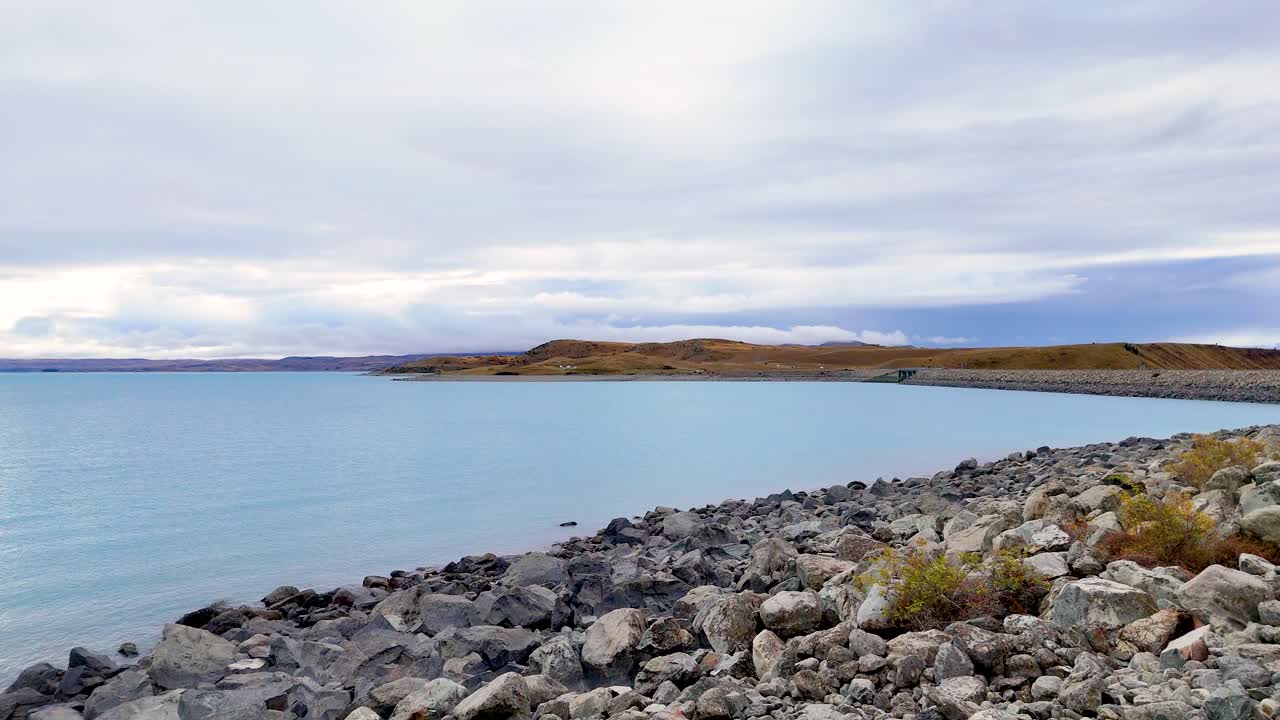 Tranquil views of Lake Pukaki's rocky shoreline under overcast skies. The camera pans smoothly, capturing the lake's calm, blue waters