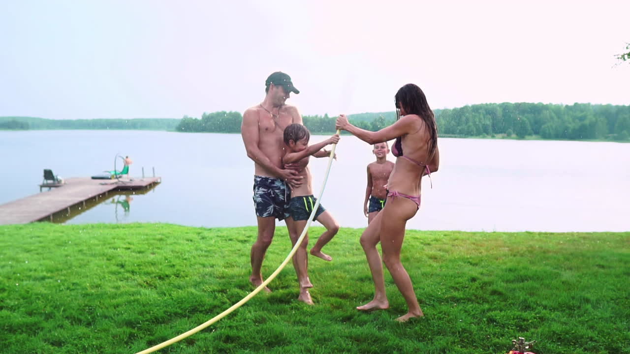 мама и двое сыновей летом на траву налили воду из шланга для газона. район загородный дом возле озера на закате