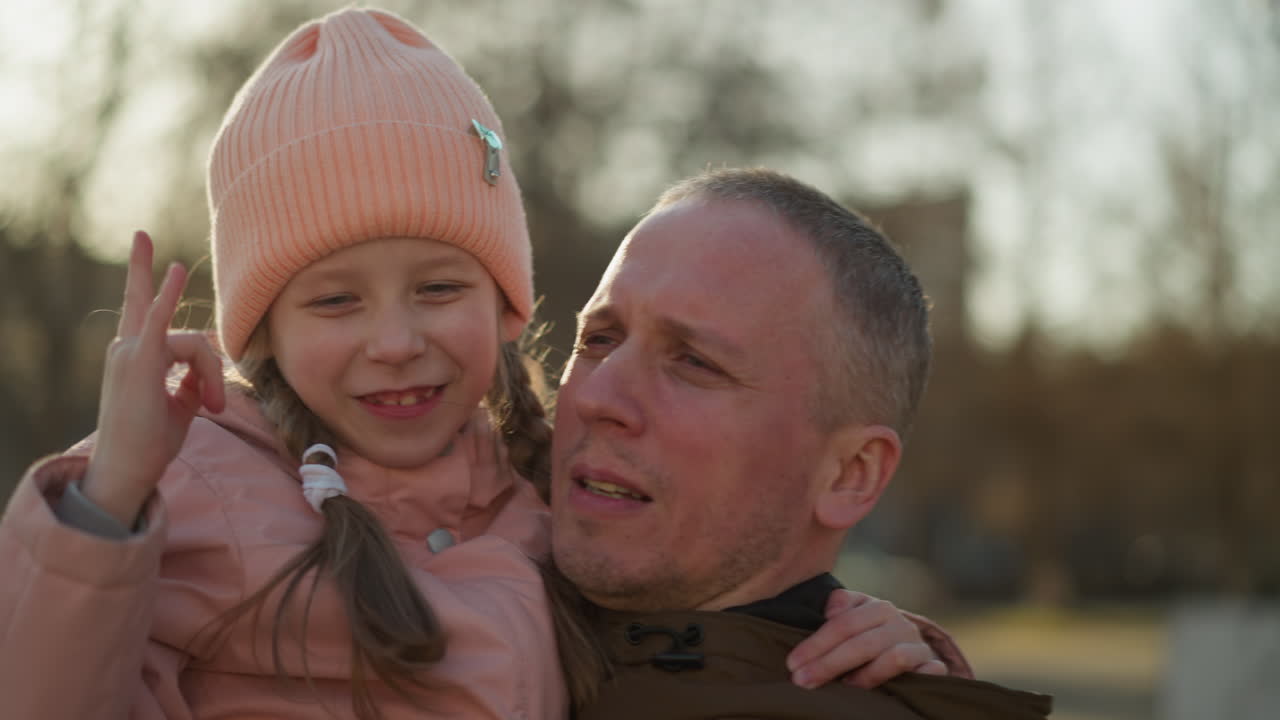 A man wearing a brown jacket carries a little girl in a pink cap and jacket as she makes a playful hand sign and they smile joyfully