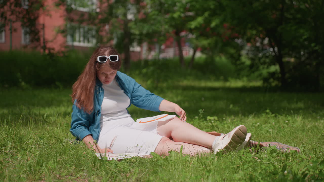 Lady sitting on blanket in sunny park comparing embroidered fabric with main design, observing details carefully, surrounded by lush green grass and trees, peaceful summer day