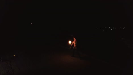 Young-blond-male-does-tricks-with-fire-spins-two-burning-torches-at-night-on-black-city-background-Travelling