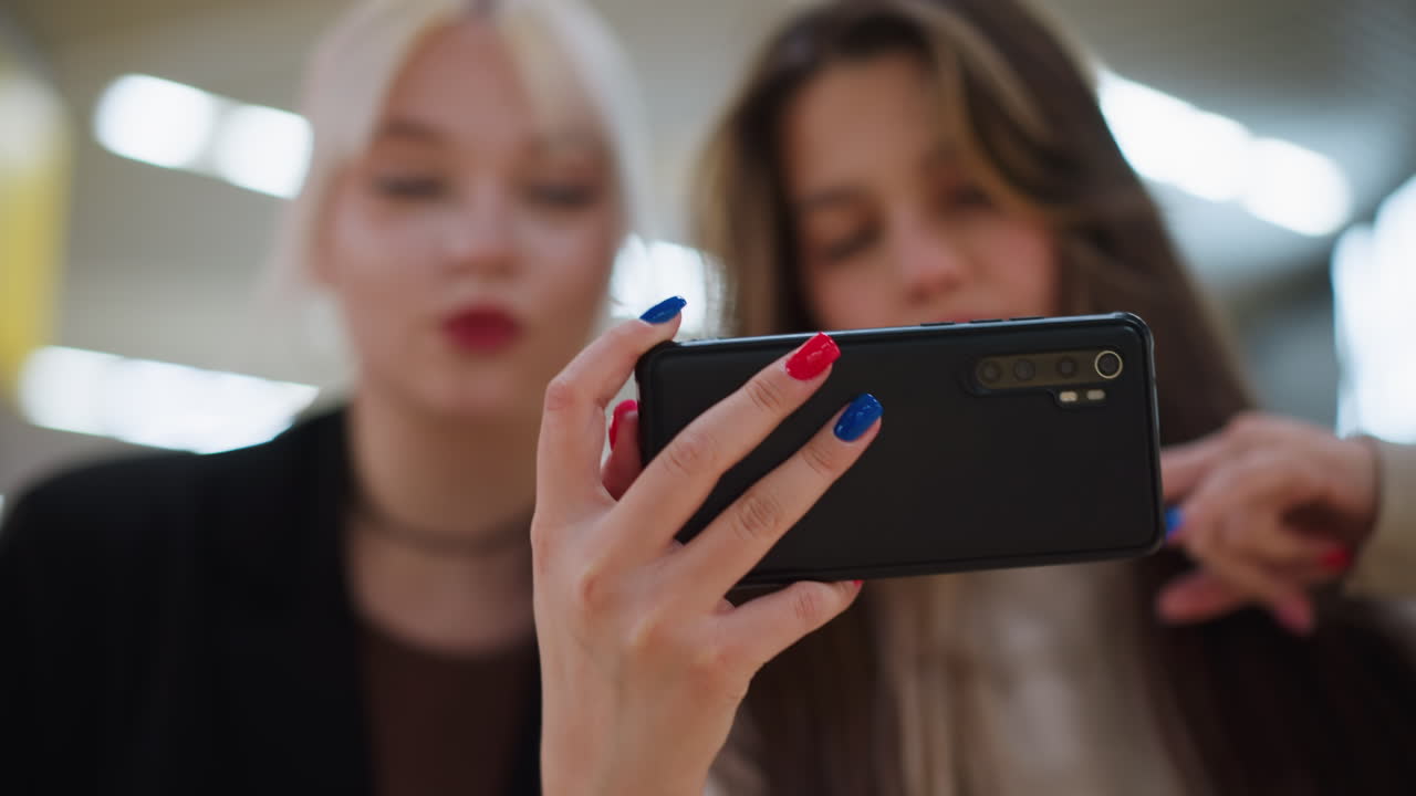Close up of ladies taking selfie with smartphone inside shopping mall, showing colorful manicured nails holding device while posing together with blurred retail environment in background