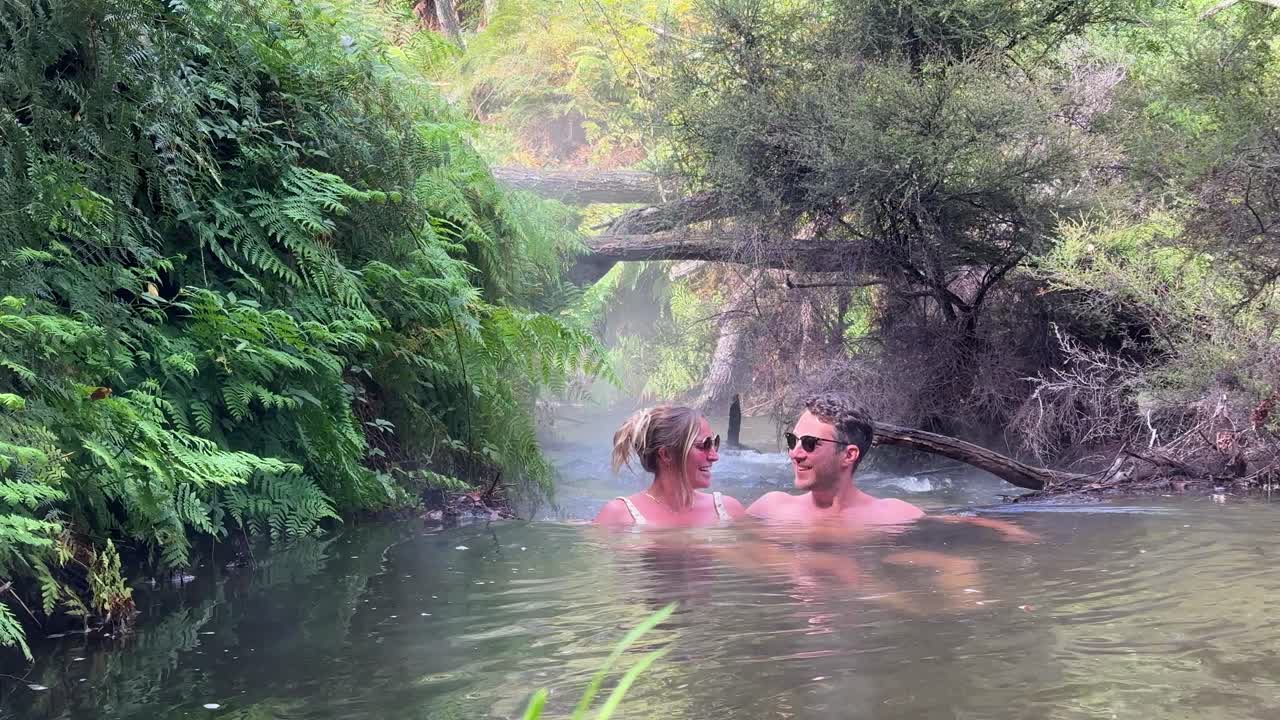 A couple relaxes in the warm, natural hot springs of Kerosene Creek, New Zealand. Surrounded by lush greenery, the steaming water creates a tranquil and peaceful scene