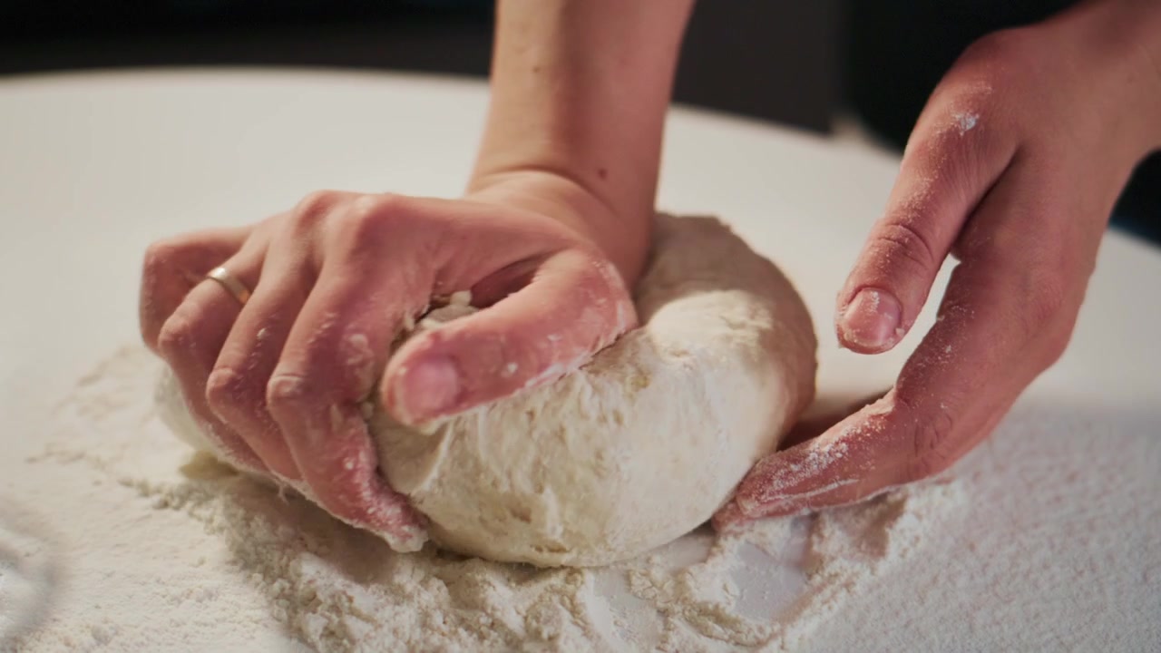 Hands knead dough on floured surface. Fingers press down, shaping soft, elastic dough. Flour dusts workspace. Focus on tactile, rhythmic motion. Ideal for recipes, baking tutorials