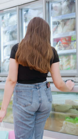 쇼핑몰에서 쇼핑하는 여성의 뒷면, 디스플레이에 멈추고, 유리 냉장고에 손을 고, 계속 걸어가고, 밝고 잘 조명된 쇼핑몰, 냉동 식품 디스플레와 활기찬 쇼핑 환경