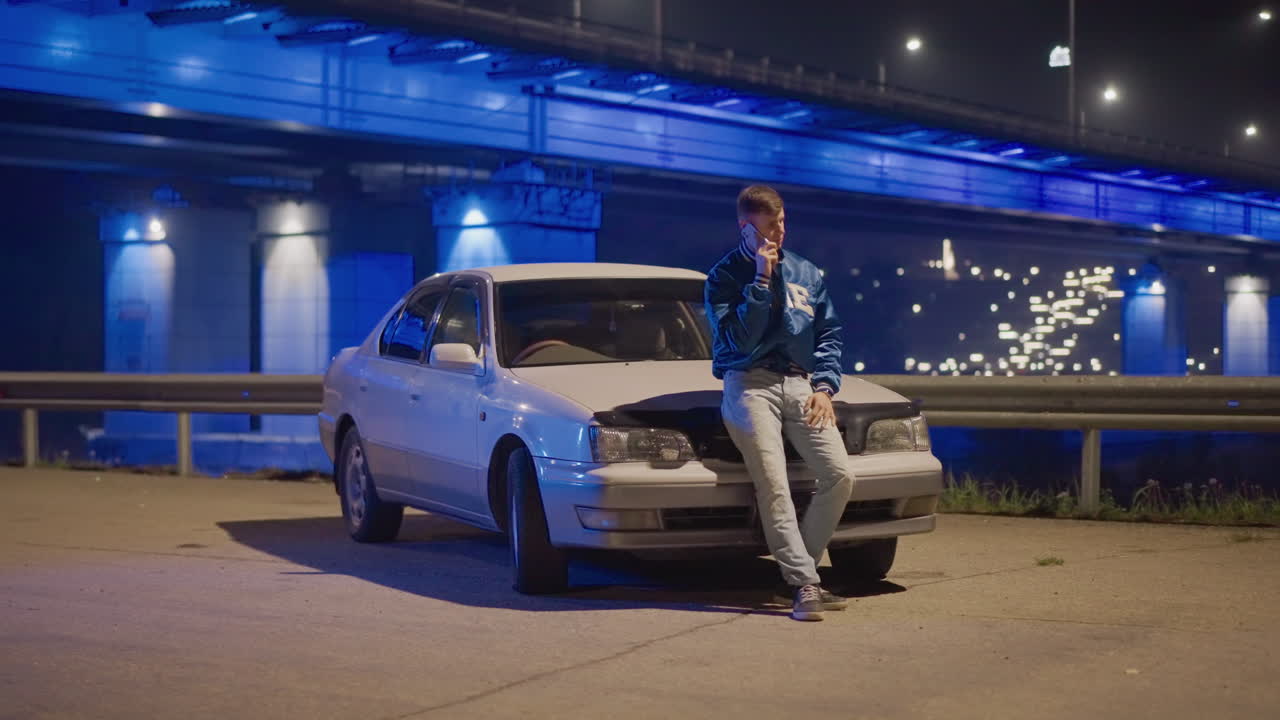 Puente azul, ciudad de noche, hombre apoyado en el capó de un coche blanco bajo luces brillantes, pose confiada con chaqueta vaquera, barandilla iluminada y farolas de la autopista, energía urbana cinematográfica y pulso nocturno
